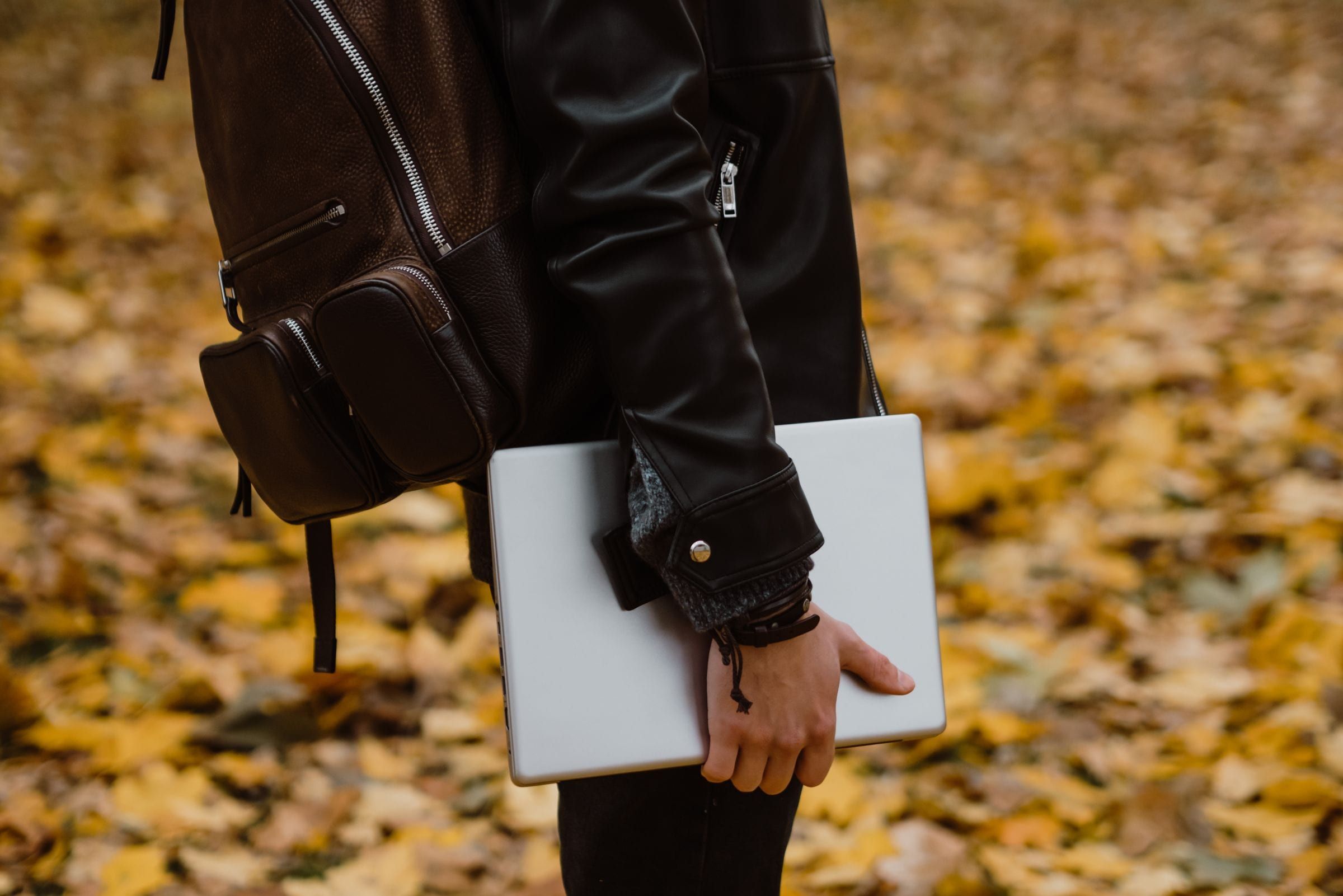 Persona con mochila y campera de cuero llevando una notebook cerrada bajo el brazo, sobre un fondo de hojas de otoño