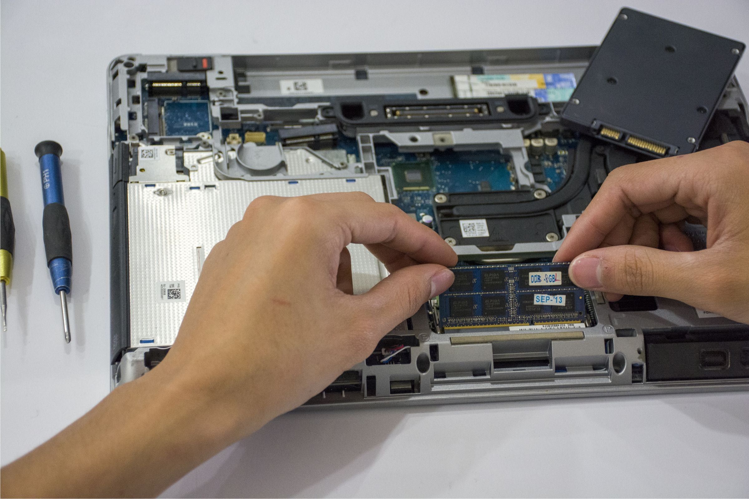 Manos colocando un módulo de memoria RAM en una notebook abierta, con un disco SSD y dos destornilladores al costado