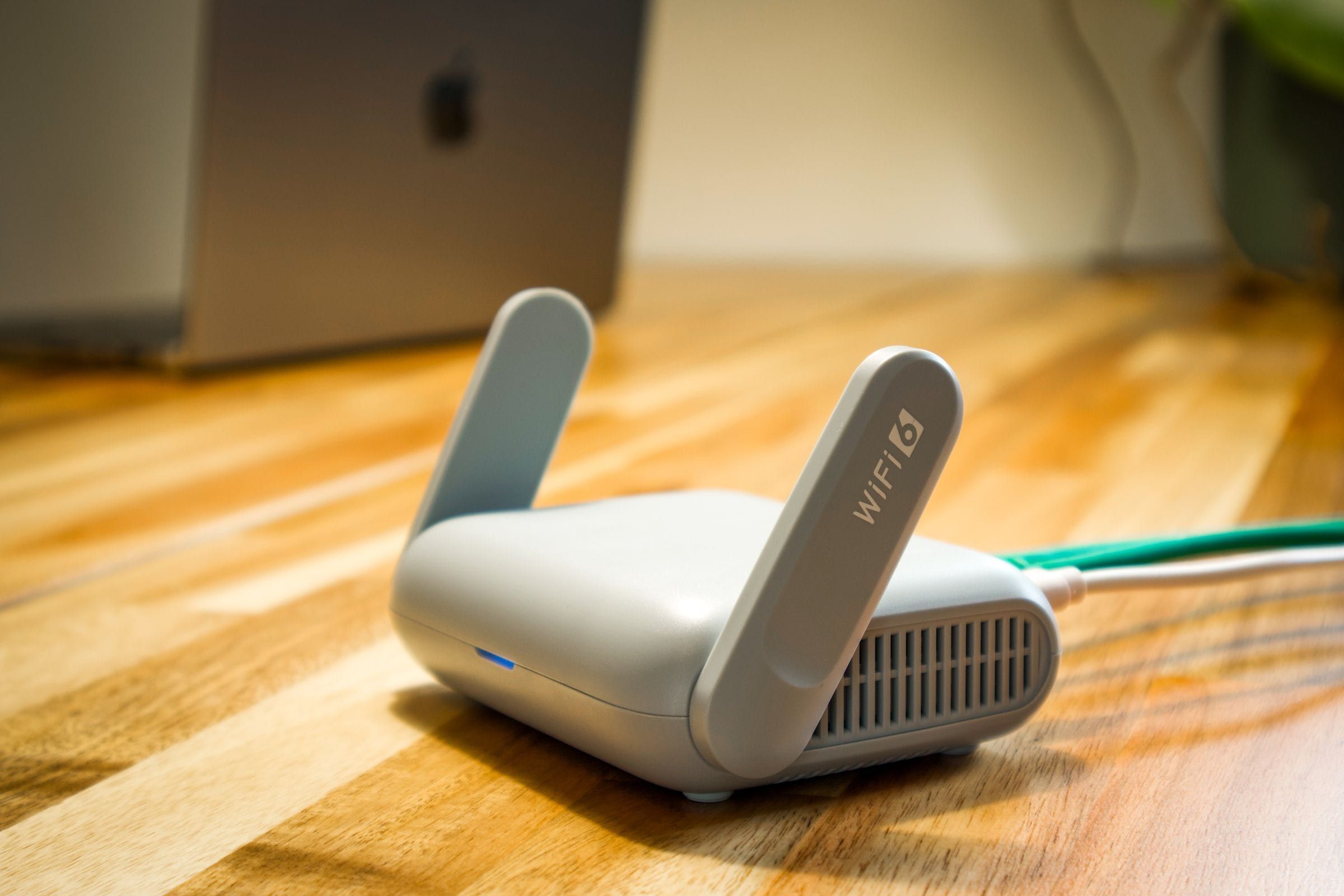 Router wifi chico con dos antenas sobre un escritorio de madera, con una notebook de fondo