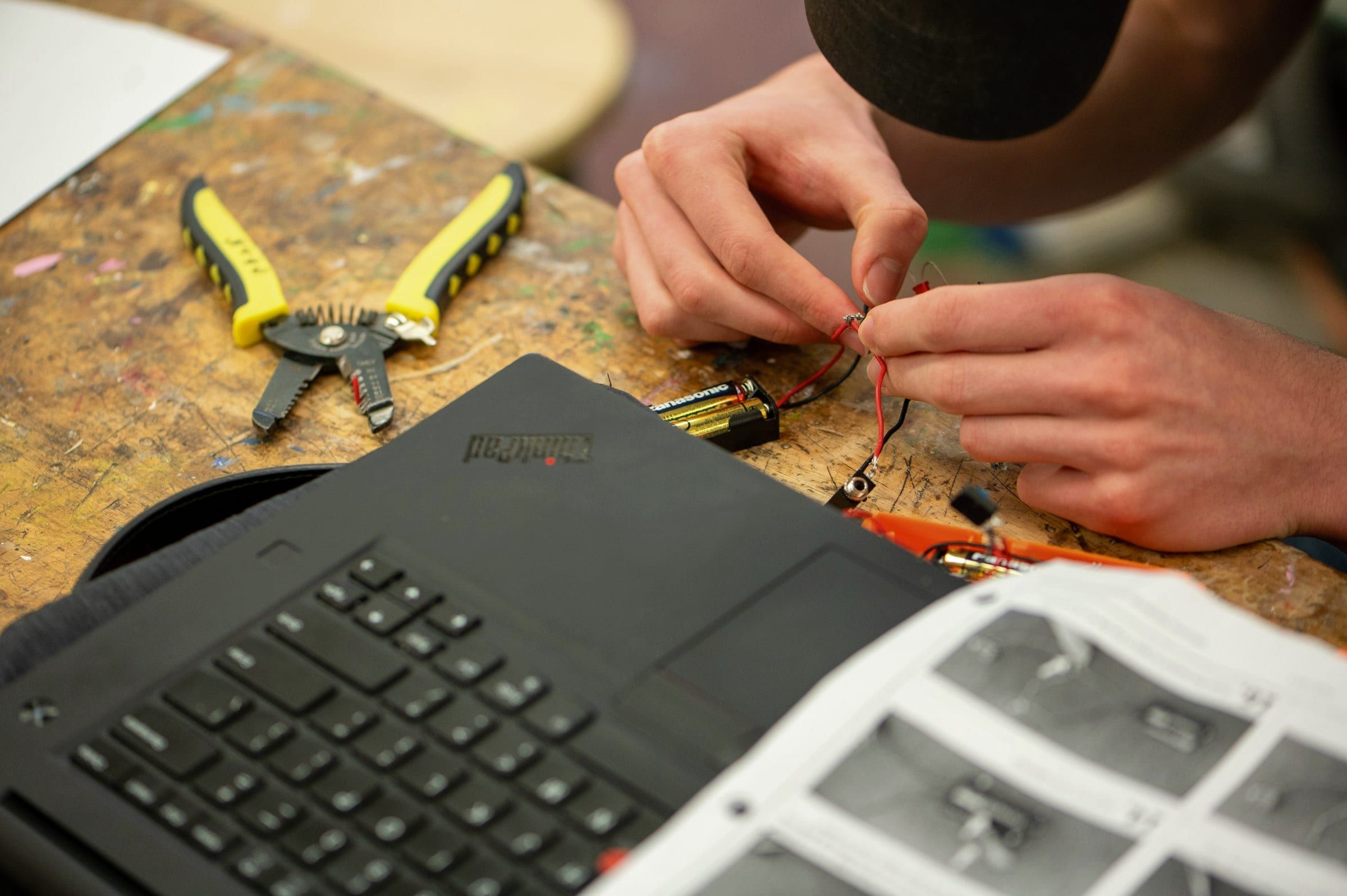 Manos de un técnico pelando y uniendo cables sobre un banco de taller, junto a una pinza amarilla y una notebook